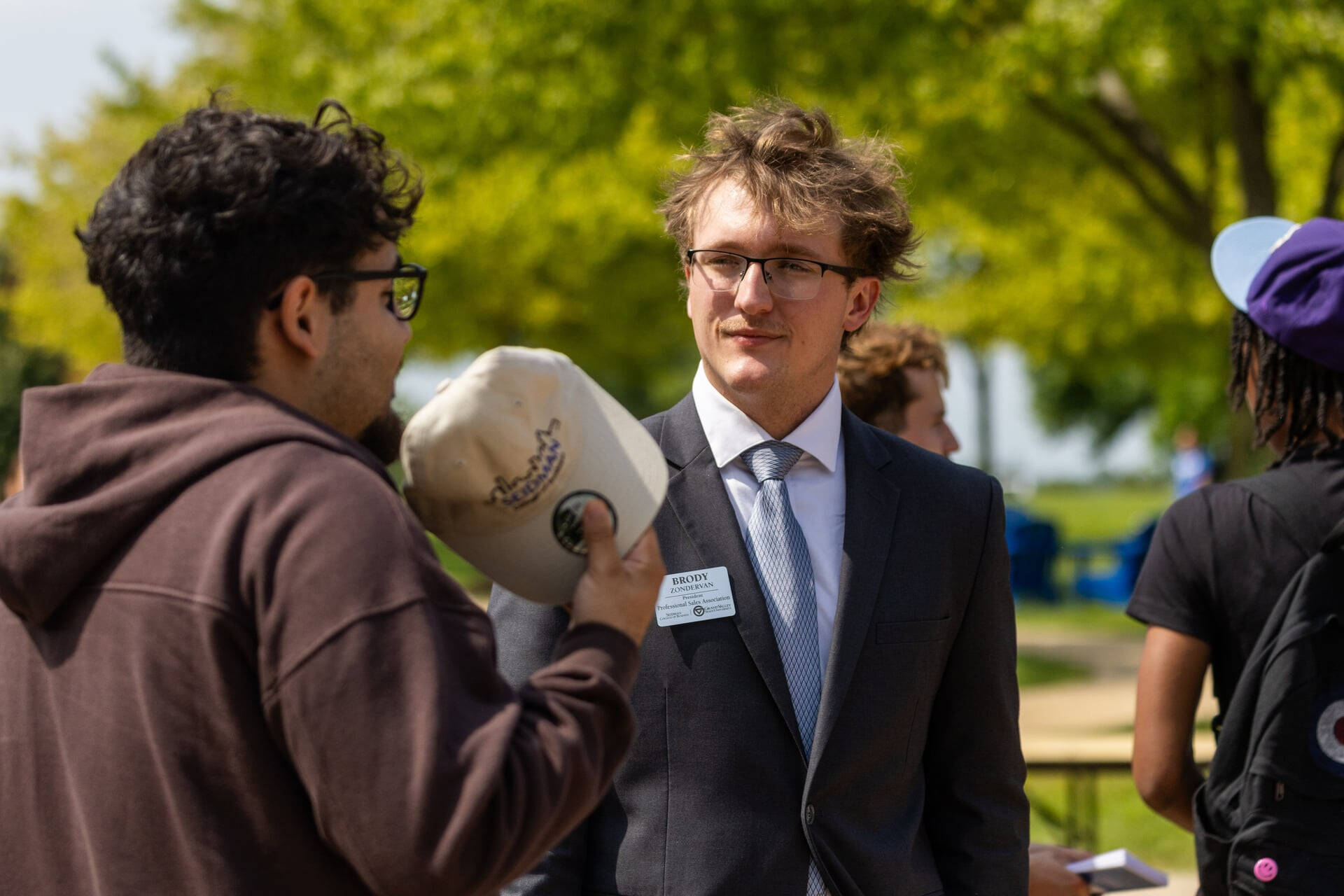 Brody Zondervan, right, in a suit jacket stands outside talking with student in brown hoodie who is holding a tan hat